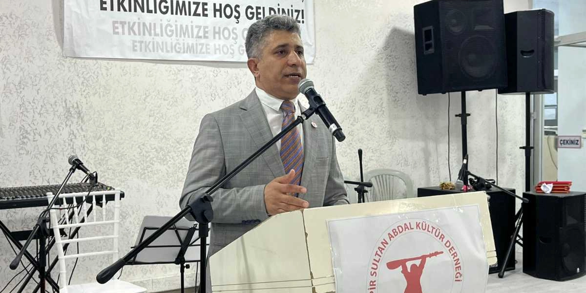 Gri takım elbiseli ve desenli kravatlı bir erkek, üzerinde Pir Sultan Abdal Kültür Derneği logosu bulunan bir kürsüde mikrofon başında konuşma yapıyor. Arka planda 