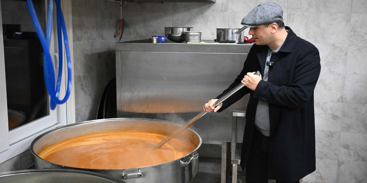 Gri kasketli ve siyah paltolu bir adam, endüstriyel bir mutfakta büyük bir metal kazanda pişmekte olan turuncu renkli çorbayı uzun bir kepçe ile karıştırıyor.