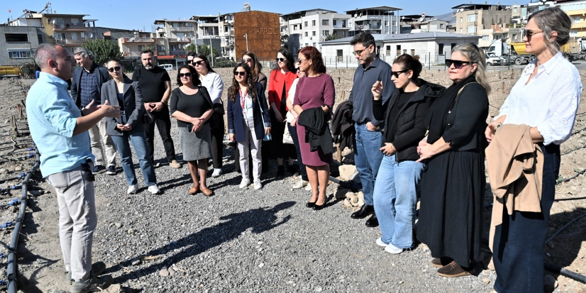 Açık havada, çakıllı bir arazide duran bir grup insan, açık mavi gömlekli bir konuşmacıyı dinliyor. Konuşmacı, sulama borularının döşendiği bir alanı işaret ederek gruba bilgi veriyor. Grubun arkasında, üzerinde 