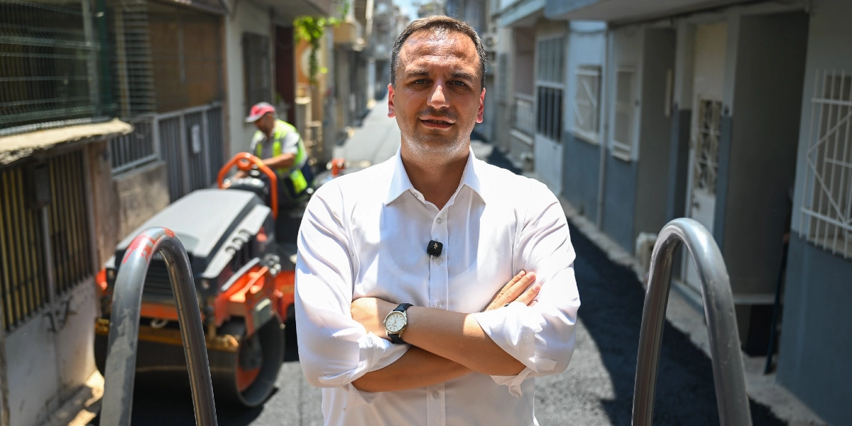 Beyaz gömlekli bir erkek, kollarını kavuşturmuş şekilde kameraya bakıyor. Arka planda dar bir sokakta asfalt serme çalışması yapılıyor ve bir iş makinesi operatörü görülüyor.