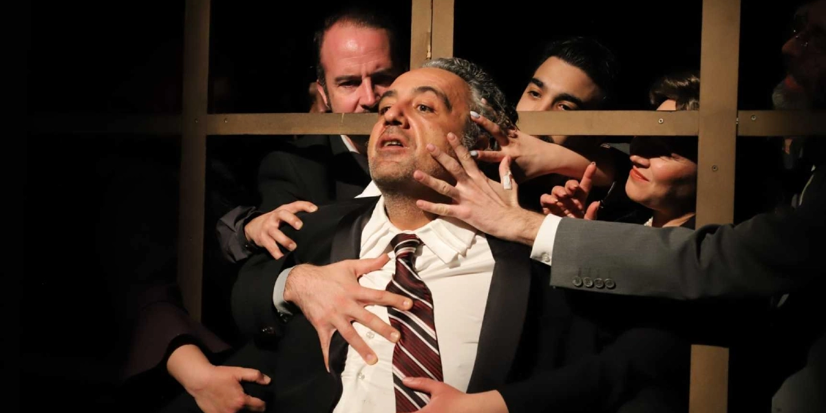 Karanlık bir sahnede, beyaz gömlek ve çizgili kravat giymiş orta yaşlı bir adamın etrafını saran oyuncular, elleriyle adamın yüzüne ve göğsüne dokunarak dramatik bir sahneyi canlandırıyor. Arka planda ahşap bir paravanın arkasından bakan oyuncuların yüzleri ve elleri, yoğun bir ifadeyle merkeze odaklanmış durumda.