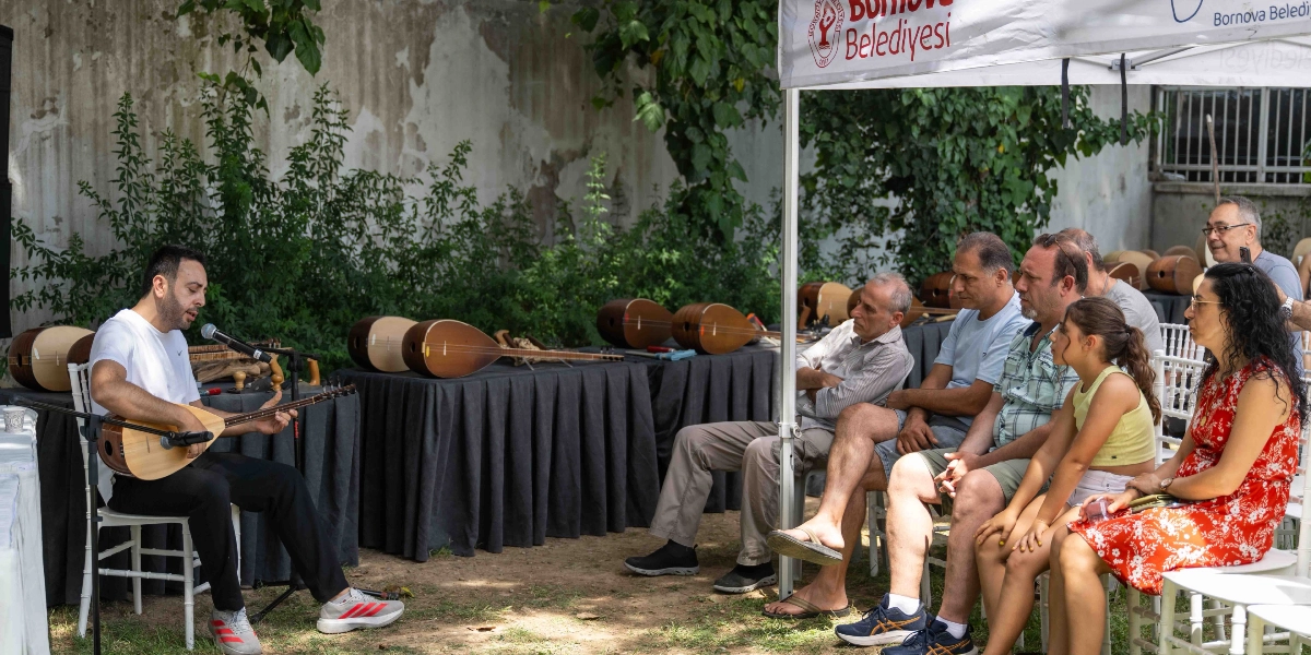 Açık havada, Bornova Belediyesi logolu bir çadırın altında düzenlenen etkinlikte, beyaz tişörtlü bir müzisyen elindeki bağlamayı çalarken izleyicilere dinleti sunuyor. Müzisyenin önündeki masalarda ve arkasındaki stantlarda yapım aşamasında olan veya tamamlanmış çok sayıda bağlama sergileniyor. İzleyiciler arasında yetişkinler ve bir çocuk yer alıyor; hepsi dikkatle müzisyeni dinliyor. Arka planda ise yeşil bitkilerle kaplı bir duvar bulunuyor.