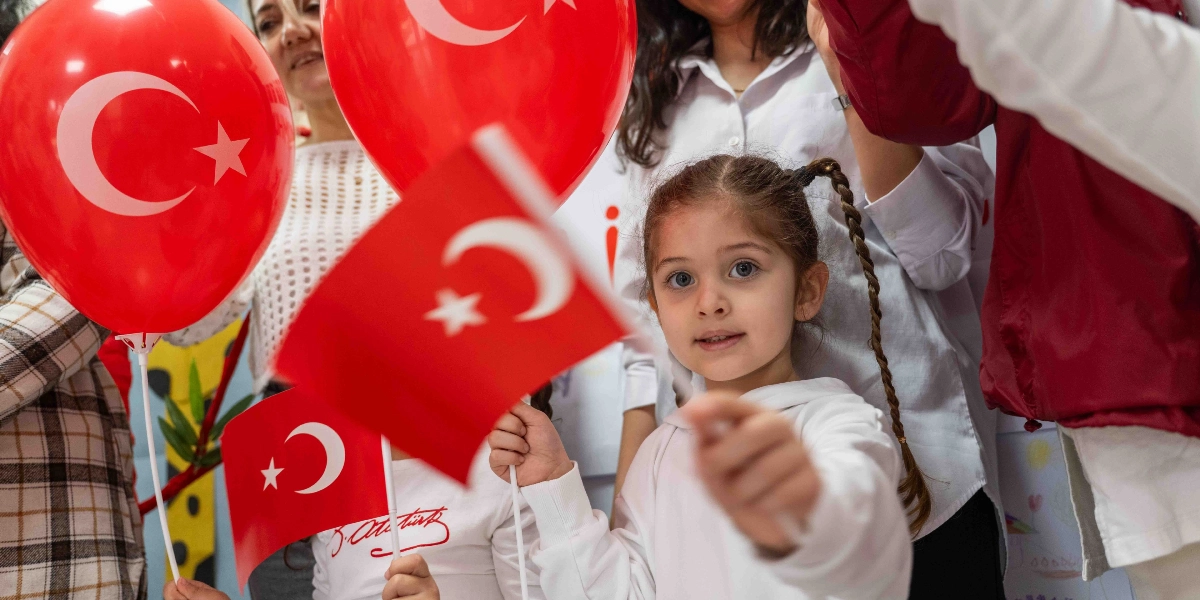 Beyaz kıyafetli, saçları örgülü küçük bir kız çocuğu, elinde tuttuğu Türk bayrağını kameraya doğru uzatıyor. Çocuğun çevresinde ellerinde üzerinde ay yıldız bulunan kırmızı balonlar ve küçük Türk bayrakları tutan diğer çocuklar ile yetişkinler yer alıyor. Ortamda neşeli bir Cumhuriyet Bayramı kutlaması havası hakim.