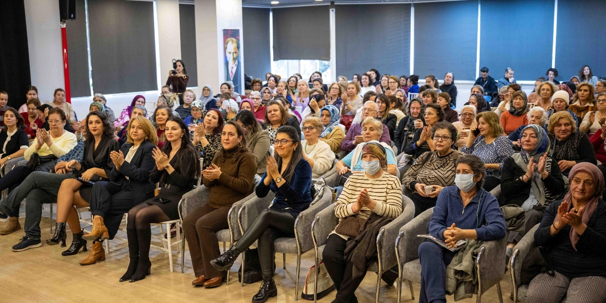 Kapalı bir etkinlik salonunda, rahim ağzı kanseri farkındalık etkinliğine katılan ve çoğunluğu kadınlardan oluşan kalabalık bir izleyici grubu, alkışlayarak bir konuşmacıyı dinliyor. Katılımcıların bir kısmı maske takarken, salonun arka duvarında Atatürk portresi ve Türk bayrağı yer alıyor.