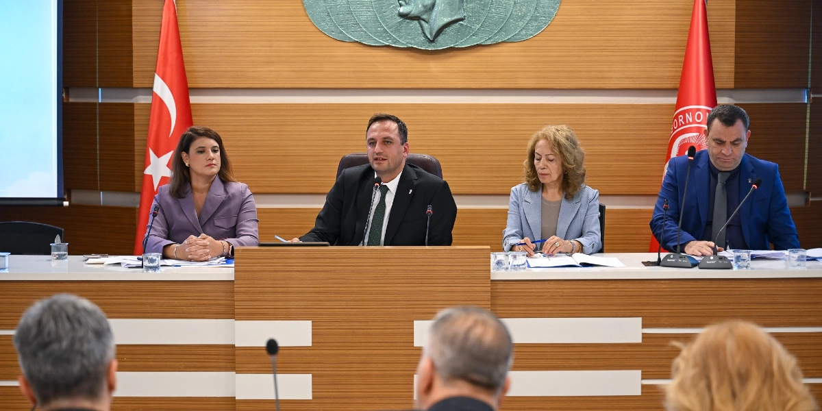 Bornova Belediyesi meclis salonunda, ahşap bir kürsüde yan yana oturan dört kişilik bir heyet toplantı yapıyor. Ortada konuşma yapan bir erkek, yanında iki kadın ve bir erkek katılımcı bulunuyor. Arka planda Türk bayrağı, Bornova Belediyesi bayrağı ve duvarda Atatürk kabartması yer alıyor. Ön planda ise toplantıyı izleyen katılımcıların başlarının arkası görülüyor.