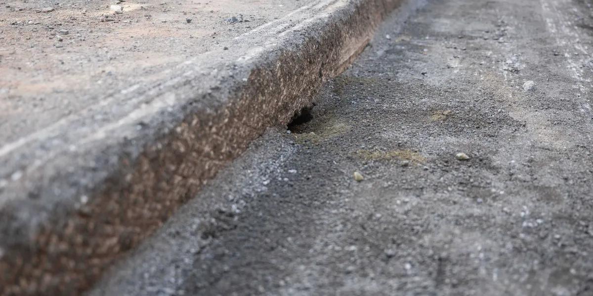 Asfaltı sökülmüş, üzerinde toprak ve çakıl kalıntıları bulunan bir yol kenarı ile kaldırım bordür taşı. Bordürün alt kısmında, zemindeki boşluktan kaynaklanan küçük bir delik görülüyor.