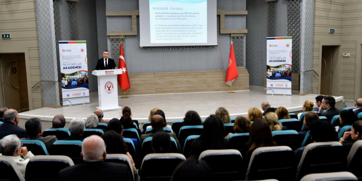 Bornova Belediyesi'nin düzenlediği Muhtarlar Akademisi etkinliğinde, kürsüde konuşma yapan bir yetkili ve onu dinleyen katılımcılar görülüyor. Salonun sahne kısmında, üzerinde Bornova Belediyesi logosu bulunan beyaz bir kürsü, iki yanında Türk bayrağı ve belediye bayrağı, arkada ise 