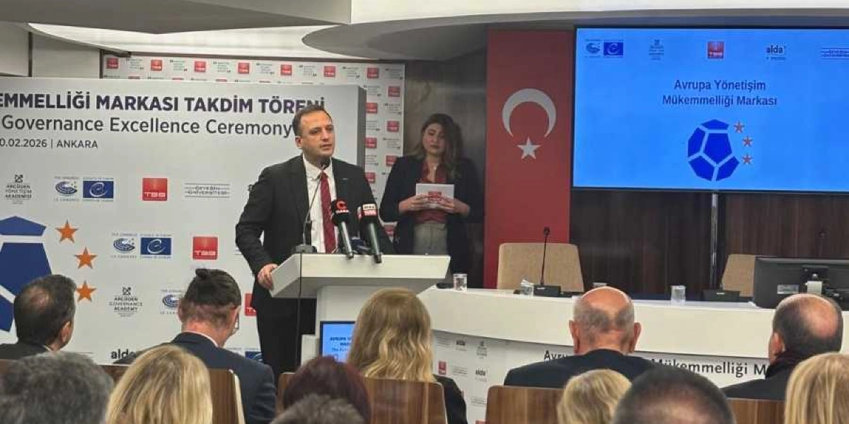 Ankara'da düzenlenen Avrupa Yönetişim Mükemmelliği Markası Takdim Töreni'nde, kürsüde konuşma yapan bir yetkili ve onu dinleyen katılımcılar yer alıyor. Arka planda Türk bayrağı ve üzerinde 