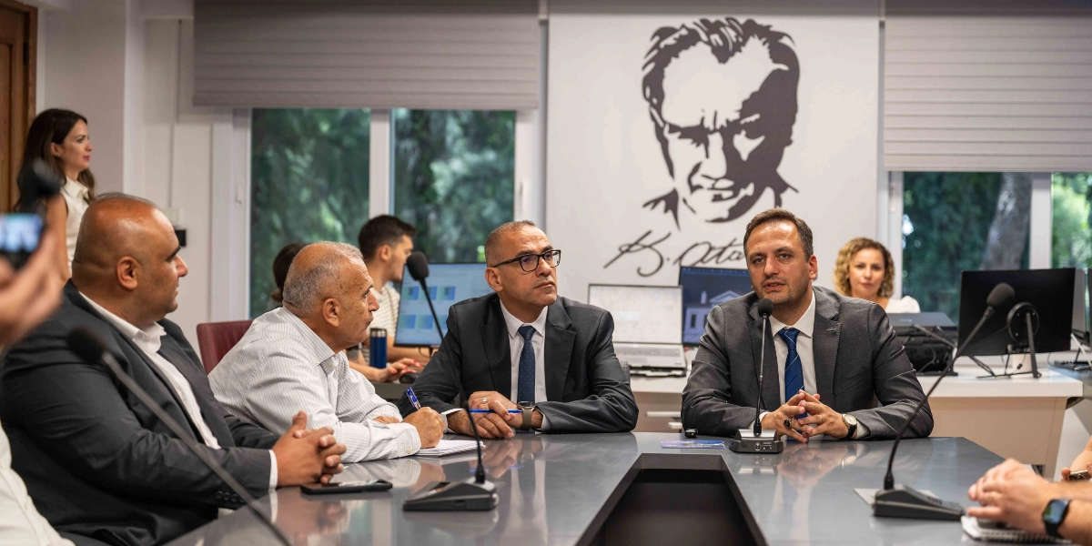 Modern bir ofis ortamında, U şeklindeki bir toplantı masasında oturan dört kişilik bir grup, önlerindeki mikrofonlarla bir toplantı veya basın açıklaması gerçekleştiriyor. Masanın başında, koyu renk takım elbiseli ve mavi kravatlı bir kişi konuşurken, yanında yine takım elbiseli ve gözlüklü bir diğer katılımcı ile açık renk gömlekli iki kişi daha oturuyor. Arka planda duvarda, Mustafa Kemal Atatürk'ün silüeti ve imzası yer alıyor. Ofis ortamında bilgisayar ekranları ve çalışma masaları görülüyor.