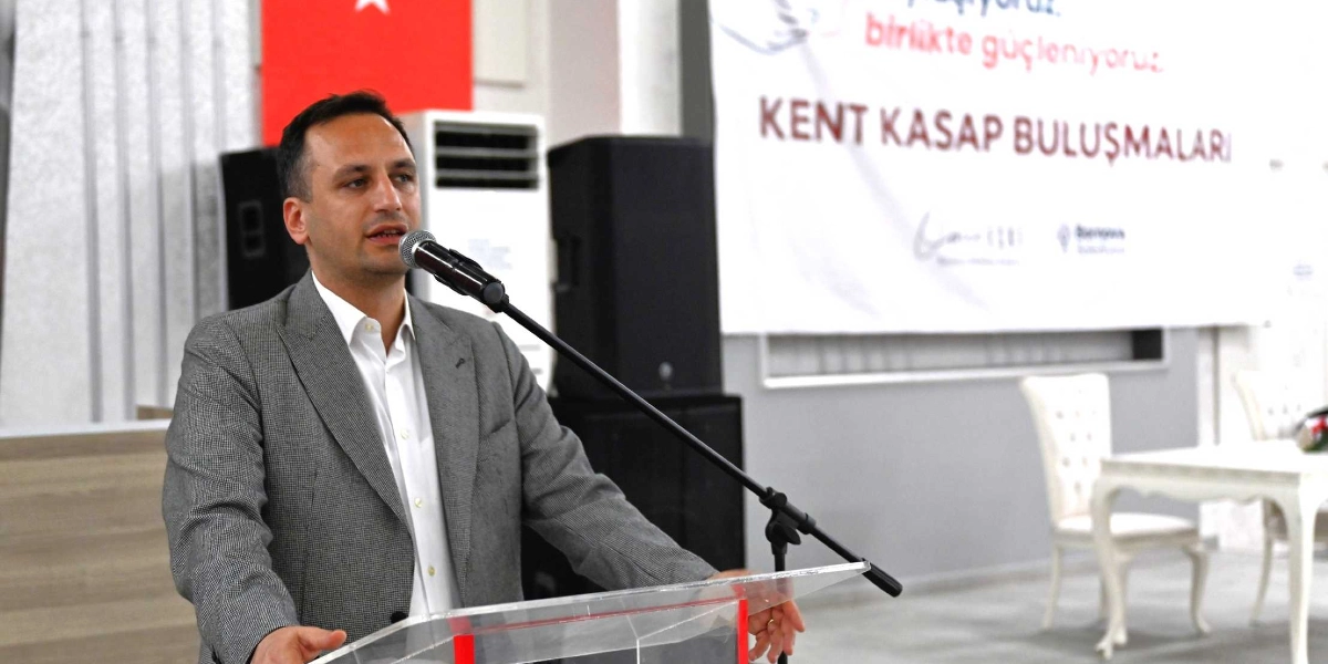 Gri ceketli ve beyaz gömlekli bir erkek, şeffaf bir kürsüde mikrofon başında konuşma yapıyor. Arka planda üzerinde 