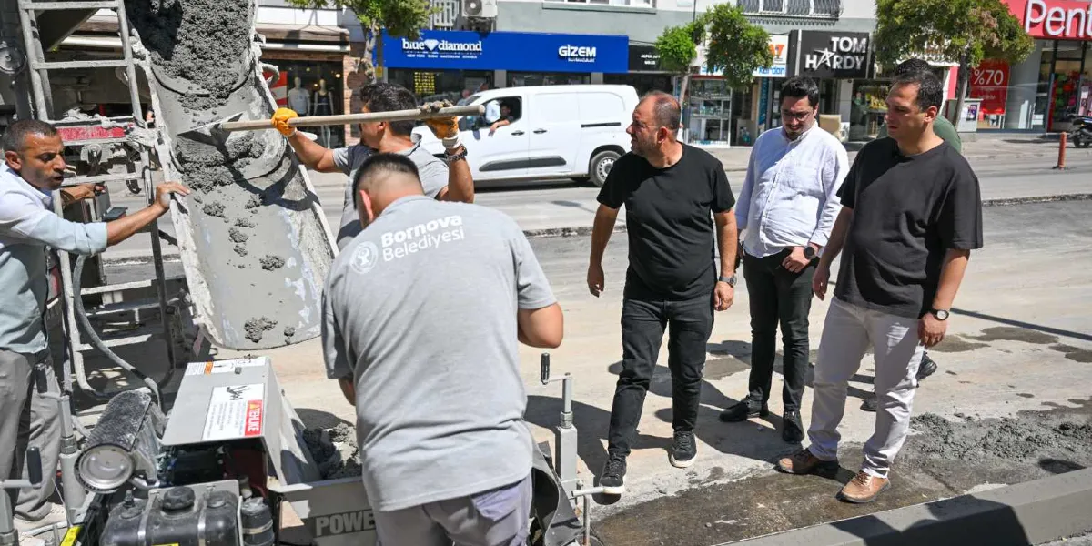Bornova Belediyesi ekipleri, bir beton mikserinden dökülen harçla kaldırım düzenleme çalışması yaparken, yanlarında duran üç yetkili çalışmaları inceleyerek bilgi alıyor. Ekiplerden birinin sırtında Bornova Belediyesi logosu bulunan gri bir tişört yer alıyor. Arka planda ise cadde üzerindeki dükkanlar ve trafik akışı görülüyor.