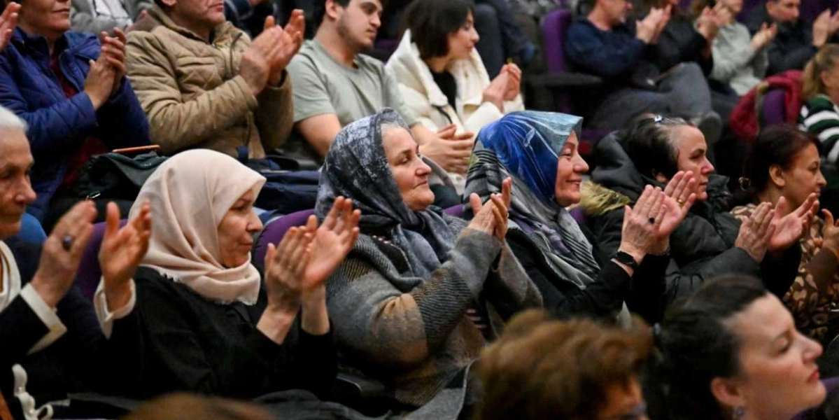 Kapalı bir salonda, mor koltuklarda oturan ve sahnedeki bir etkinliği izleyerek alkışlayan çeşitli yaş ve giyim tarzlarından oluşan kalabalık bir izleyici grubu. Ön planda başörtülü kadınların ağırlıkta olduğu izleyiciler, dikkatle sahneye bakarak elleriyle alkış tutuyor.