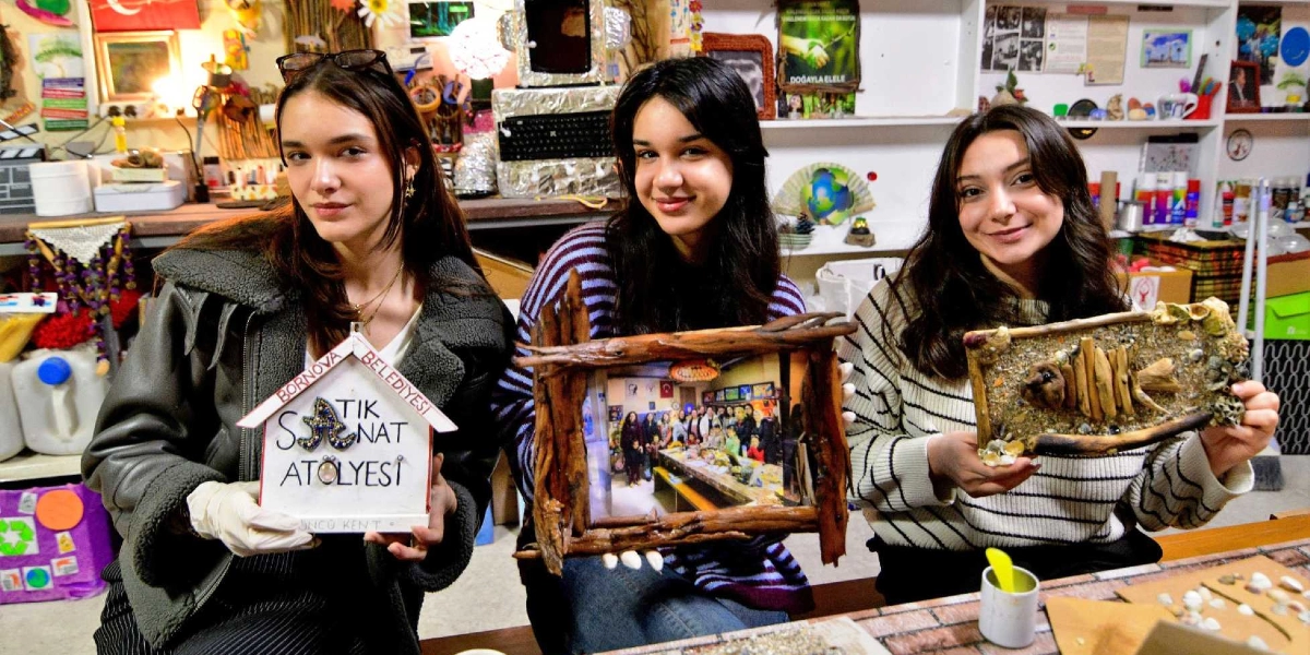 Bornova Belediyesi Atık Sanat Atölyesi'nde üç genç kadın, atık malzemelerden yaptıkları sanat eserlerini ellerinde tutarak gülümsüyor. Soldaki genç kadın üzerinde 