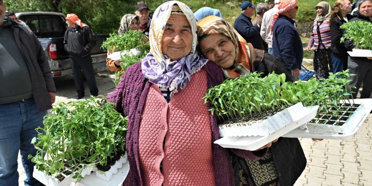 Başörtülü iki yaşlı kadın, ellerinde tuttukları plastik viyollerdeki yeşil sebze fideleriyle gülümsüyor. Arka planda, açık havada düzenlenen bir dağıtım etkinliğinde bekleyen diğer insanlar ve bir kamyonet görülüyor.