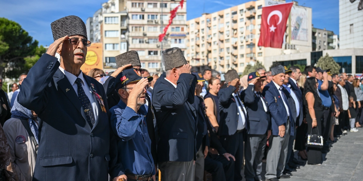 Bornova'da düzenlenen 19 Eylül Gaziler Günü töreninde, resmi kıyafetli ve kalpaklı gaziler ile vatandaşlar saygı duruşunda bulunuyor. Ön planda, lacivert takım elbiseli ve kalpaklı bir gazi ile yanında lacivert gömlekli ve üzerinde Türk bayrağı amblemli şapka bulunan bir başka gazi asker selamı veriyor. Arka planda ise törene katılan diğer gaziler, sivil vatandaşlar ve binaların arasında asılı duran büyük bir Türk bayrağı görülüyor.