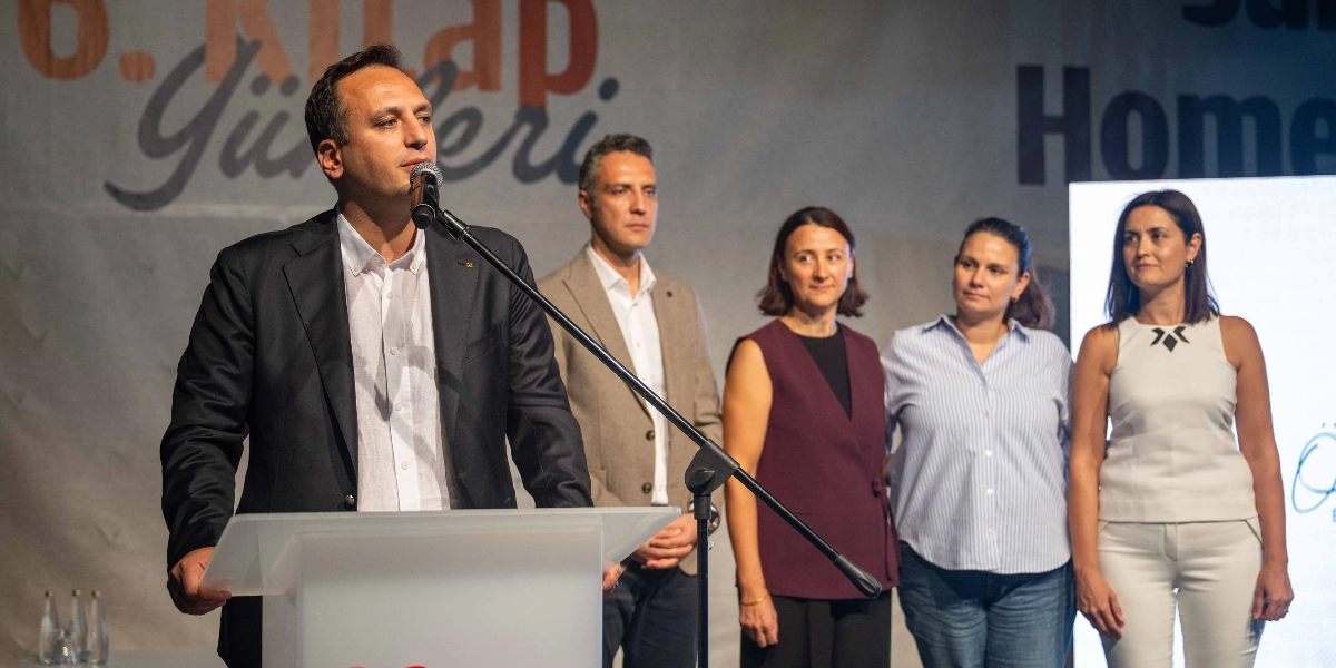Bornova Kitap Günleri etkinliğinde kürsüde konuşma yapan bir erkek ve onu dinleyen dört kişilik bir grup. Konuşmacı koyu renkli bir ceket ve beyaz gömlek giymiş, mikrofon başında duruyor. Arka planda 