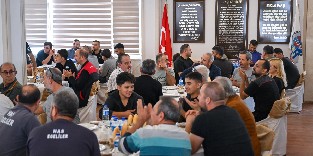 3. Sanayi Sitesi'nde düzenlenen bir yemekte, beyaz örtülü masaların etrafında toplanmış çok sayıda erkek katılımcı bir arada oturuyor. Bazı katılımcılar gülümseyerek sohbet ediyor, bazıları ise ellerini çırparak alkışlıyor. Masaların üzerinde su şişeleri ve ekmekler bulunuyor. Arka planda duvarda asılı Türk bayrağı, Gençliğe Hitabe ve İstiklal Marşı metinlerinin yer aldığı çerçeveli panolar ile sanayi sitesine ait bir logo dikkat çekiyor.