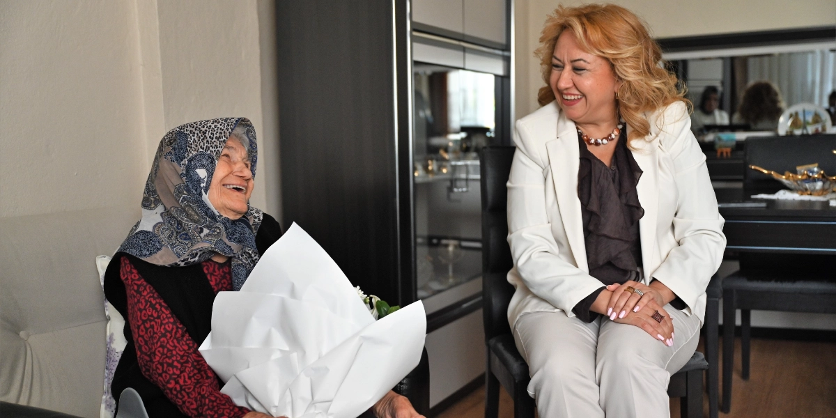 Beyaz ceketli sarışın bir kadın, ev ortamında oturan yaşlı bir kadını ziyaret ederek onunla neşeyle sohbet ediyor. Yaşlı kadın başında desenli bir eşarp ile kucağında beyaz ambalajlı bir çiçek buketi tutuyor ve gülümseyerek ziyaretçisine bakıyor.