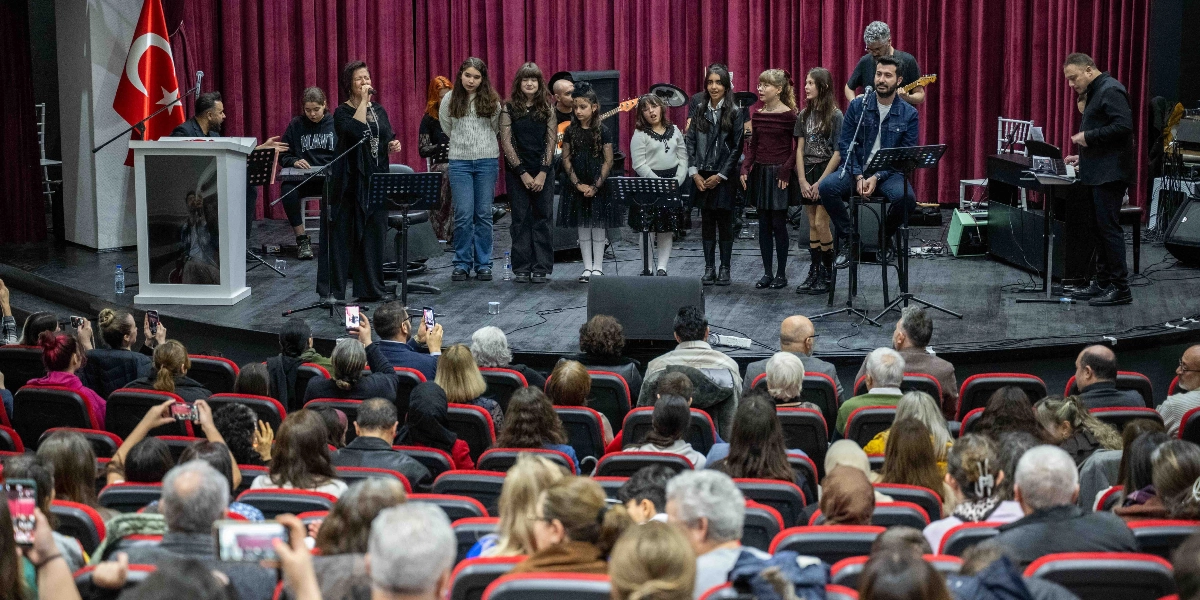 Kırmızı perdeli bir sahnede, mikrofon başında şarkı söyleyen bir kadın solist ve yanında duran gençlerden oluşan bir koro grubu yer alıyor. Sahnenin sağ tarafında bir erkek şarkıcı taburede oturarak eşlik ediyor, arkalarında ise müzisyenler ve enstrümanlar bulunuyor. Sahnenin sol köşesinde Türk bayrağı asılı. İzleyicilerle dolu bir salonda, birçok kişi telefonlarıyla sahneyi kaydediyor veya fotoğraf çekiyor.