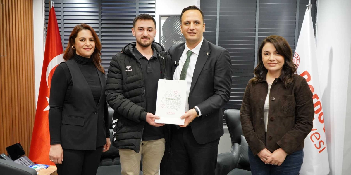 Ofis ortamında, Bornova Belediye Başkanı olduğu anlaşılan takım elbiseli ve yeşil kravatlı bir erkek, genç bir erkeğe üzerinde Bornova Belediyesi logosu ve çeşitli simgeler bulunan beyaz bir hediye çantası takdim ediyor. Yanlarında biri siyah yelekli, diğeri kahverengi ceketli iki kadın yer alıyor. Arka planda Türk bayrağı ve Bornova Belediyesi bayrağı bulunuyor.