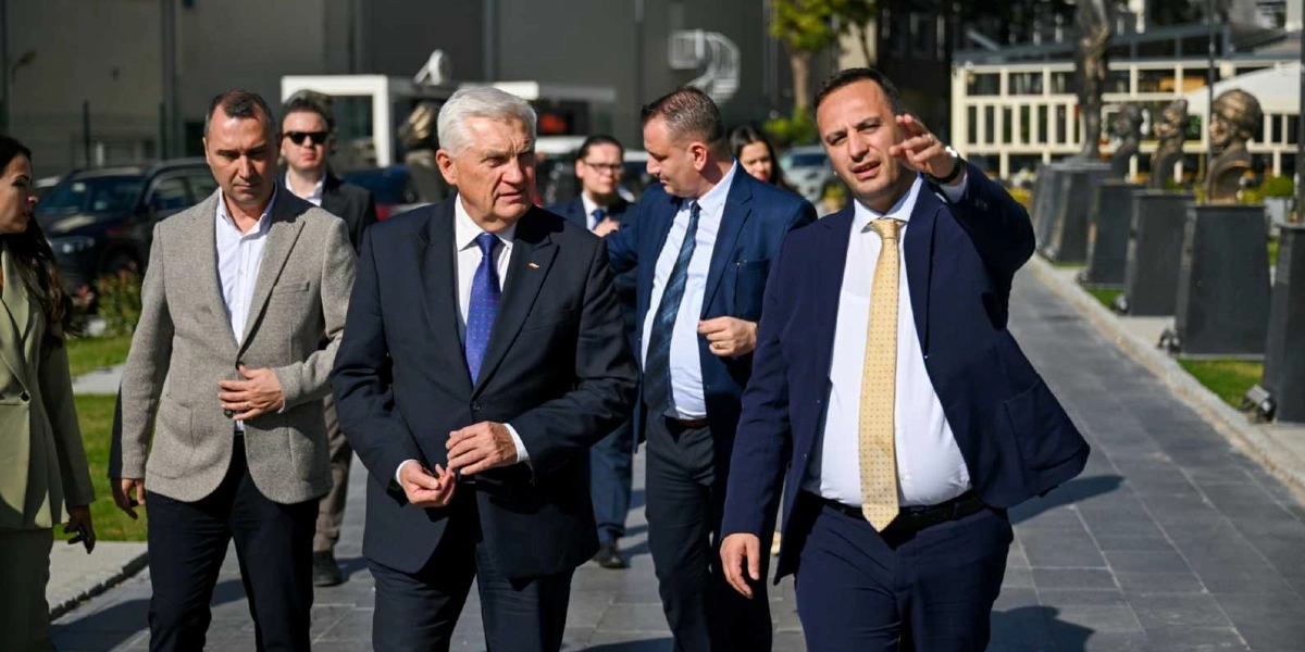 Resmi kıyafetli bir grup insan, açık havada yürürken bir yetkili tarafından çevredeki büstlerin bulunduğu alana doğru yönlendiriliyor. Ön planda, lacivert takım elbiseli ve sarı desenli kravatlı bir kişi elini ileriye doğru uzatarak bir şeyler anlatırken, yanında koyu renk takım elbiseli ve mavi kravatlı, gri saçlı bir beyefendi ile bej ceketli bir başka kişi ona eşlik ediyor. Arka planda ise yürüyüş yolunun kenarında sıralanmış büstler ve modern bir yapı görülüyor.