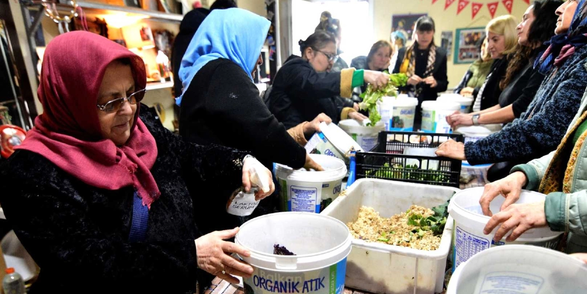 Kapalı bir mekanda bir araya gelen kadınlar, önlerindeki plastik kovalara organik atıkları yerleştirerek kompost hazırlıyor. Ön planda kırmızı başörtülü bir kadın, elindeki sprey şişesiyle kovaya sıvı püskürtüyor. Yanındaki diğer kadınlar ise ellerindeki yeşil yapraklı sebze atıklarını ve diğer organik malzemeleri beyaz kovalara dolduruyor. Masanın üzerinde 