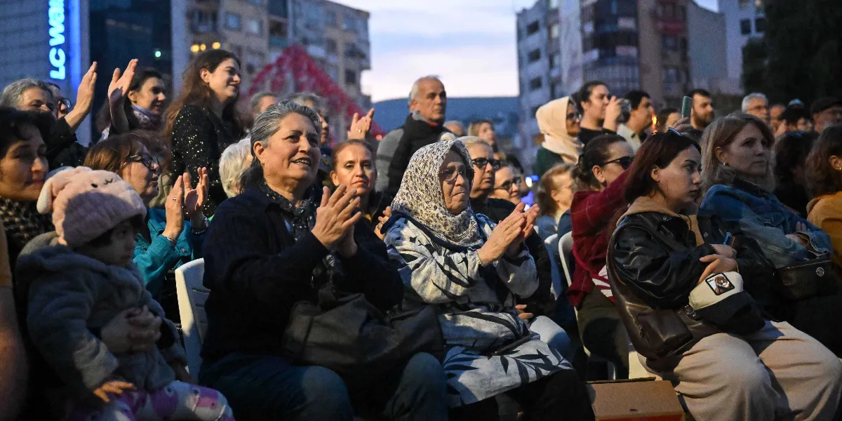 Açık havada düzenlenen bir etkinlikte, sandalyelerde oturarak alkışlayan ve sahneyi izleyen farklı yaş gruplarından oluşan kalabalık bir izleyici grubu. Ön planda kucağında küçük bir çocuk tutan bir kadın, yanında gri desenli bir başörtüsü takan bir kadın ve diğer izleyiciler yer alıyor. Arka planda ise şehir merkezindeki binalar ve akşamüstü ışığı görülüyor.