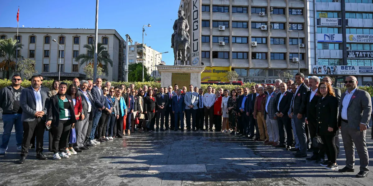Bornova Cumhuriyet Meydanı'nda, Atatürk anıtının önünde yan yana dizilmiş, çoğunluğu takım elbiseli erkekler ve şık giyimli kadınlardan oluşan kalabalık bir grup, toplu fotoğraf çekimi için kameraya bakıyor. Arka planda Bornova Hükümet Konağı binası ve üzerinde çeşitli iş yeri tabelalarının bulunduğu çok katlı bir yapı yer alıyor.