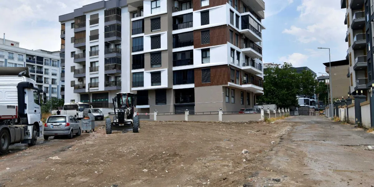Modern apartman binalarının önündeki toprak zeminli alanda, bir iş makinesi ve park halindeki araçların bulunduğu çalışma sahası.