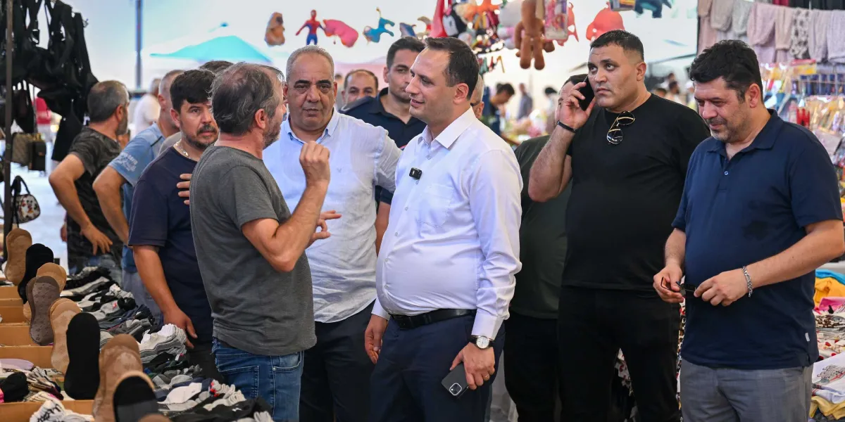 Bornova Belediye Başkanı Ömer Eşki, bir pazar yerinde esnaflarla bir araya gelerek incelemelerde bulunuyor. Beyaz gömlekli ve lacivert pantolonlu olan Başkan Eşki, pazar tezgahlarının önünde duran bir esnafla sohbet ederken, çevresinde diğer esnaflar ve belediye yetkilileri yer alıyor. Arka planda çeşitli ürünlerin sergilendiği pazar tezgahları ve oyuncaklar görülüyor.