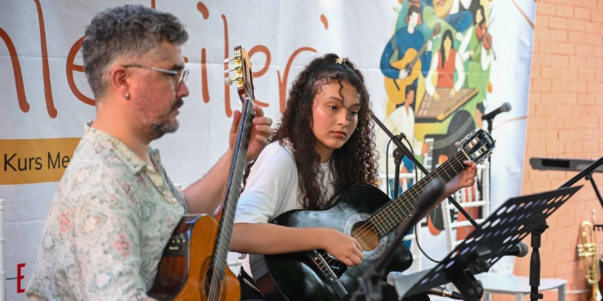 Gözlüklü, sakallı bir erkek ve uzun kıvırcık saçlı genç bir kadın, arkalarında müzik temalı illüstrasyonların yer aldığı bir afişin önünde yan yana oturarak klasik gitar çalıyor. Önlerinde nota sehpaları ve mikrofonlar bulunuyor.