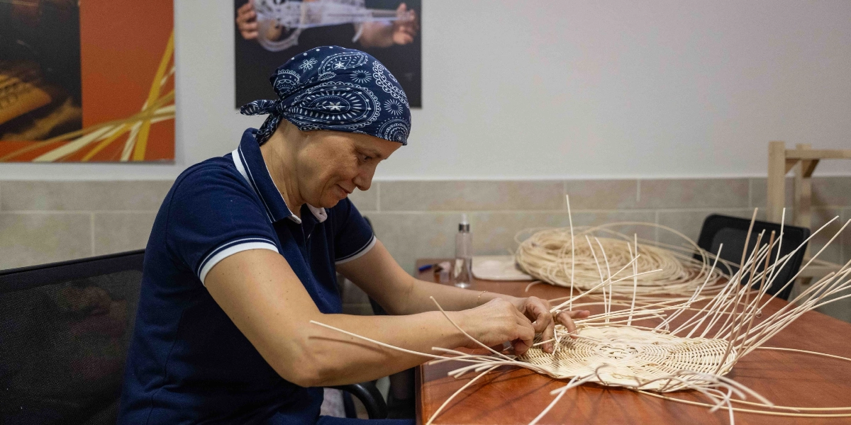 Lacivert bandana takan bir kadın, ahşap bir masanın başında oturmuş, ince söğüt dallarını kullanarak sepet örme işlemiyle uğraşıyor. Kadın, odaklanmış ve huzurlu bir ifadeyle elindeki örgüye şekil verirken, masanın üzerinde henüz tamamlanmamış sepet ve bir miktar daha malzeme bulunuyor.