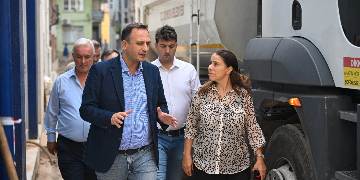 Bornova Belediyesi'ne ait bir iş makinesinin yanında yürüyen, aralarında Bornova Belediye Başkanı Mustafa İduğ'un da bulunduğu bir grup yetkili, devam eden yol çalışmalarını inceliyor. Başkan İduğ, konuşarak çevresindekilere bilgi verirken, yanındaki kadın ve diğer yetkililer onu dinliyor. İş makinesinin üzerinde 