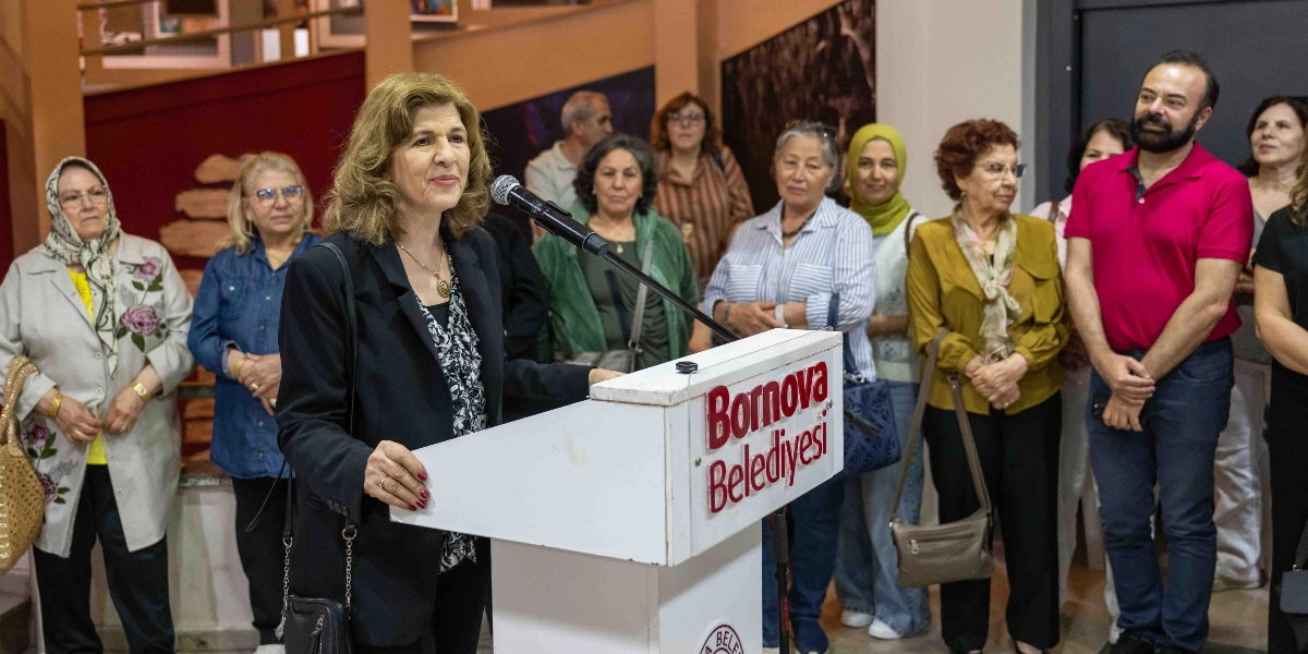 Bornova Belediyesi kürsüsünde konuşma yapan orta yaşlı bir kadın ve onu dinleyen, aralarında farklı yaş ve giyim tarzlarına sahip kadın ve erkeklerin bulunduğu bir grup sanatsever.