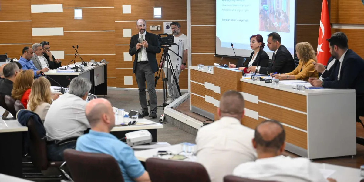 Modern bir toplantı salonunda, kürsüde elinde mikrofonla konuşma yapan bir erkek ve onu dinleyen katılımcılar yer alıyor. Salonun ön kısmındaki masada dört kişilik bir heyet oturuyor; arkalarındaki dev ekranda geri dönüşüm tesisiyle ilgili sunum yansıtılıyor. Salonun arka tarafında bir kameraman çekim yaparken, diğer katılımcılar masalarda oturarak sunumu takip ediyor. Köşede ise bir Türk bayrağı bulunuyor.