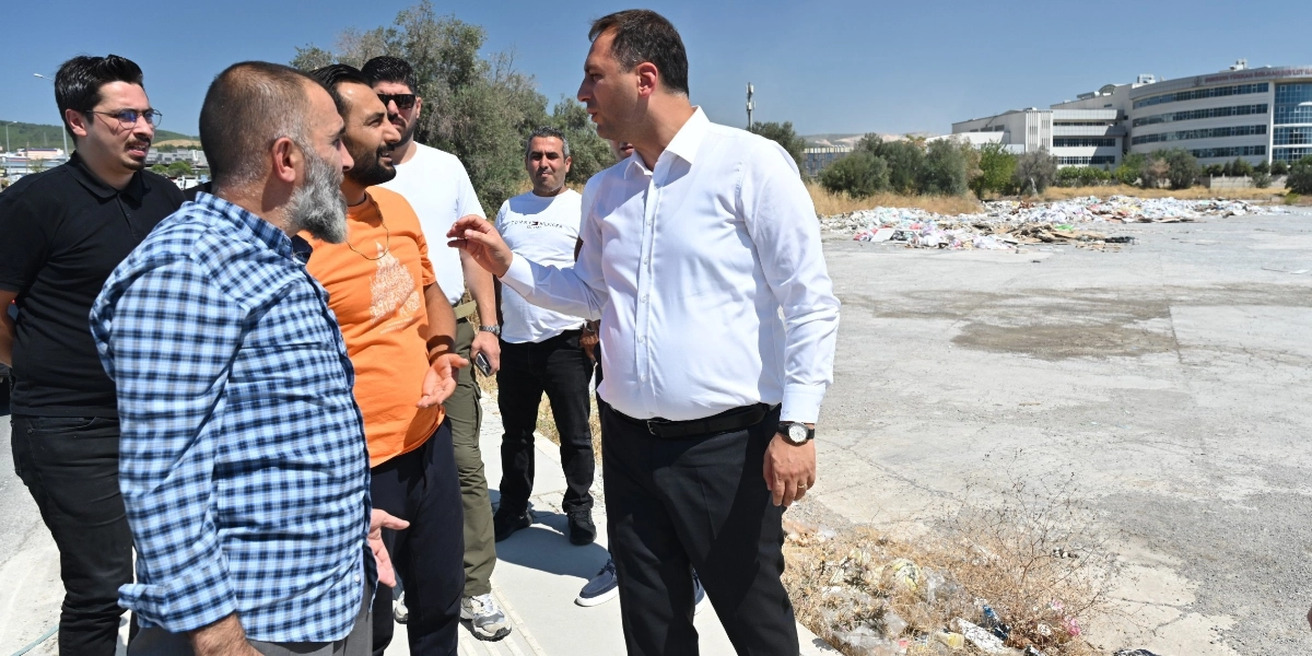 Beyaz gömlekli bir yetkili, yanında bulunan bir grup erkekle birlikte, arkada moloz yığınlarının bulunduğu boş bir arazide incelemelerde bulunuyor ve eliyle alanı işaret ederek açıklama yapıyor.