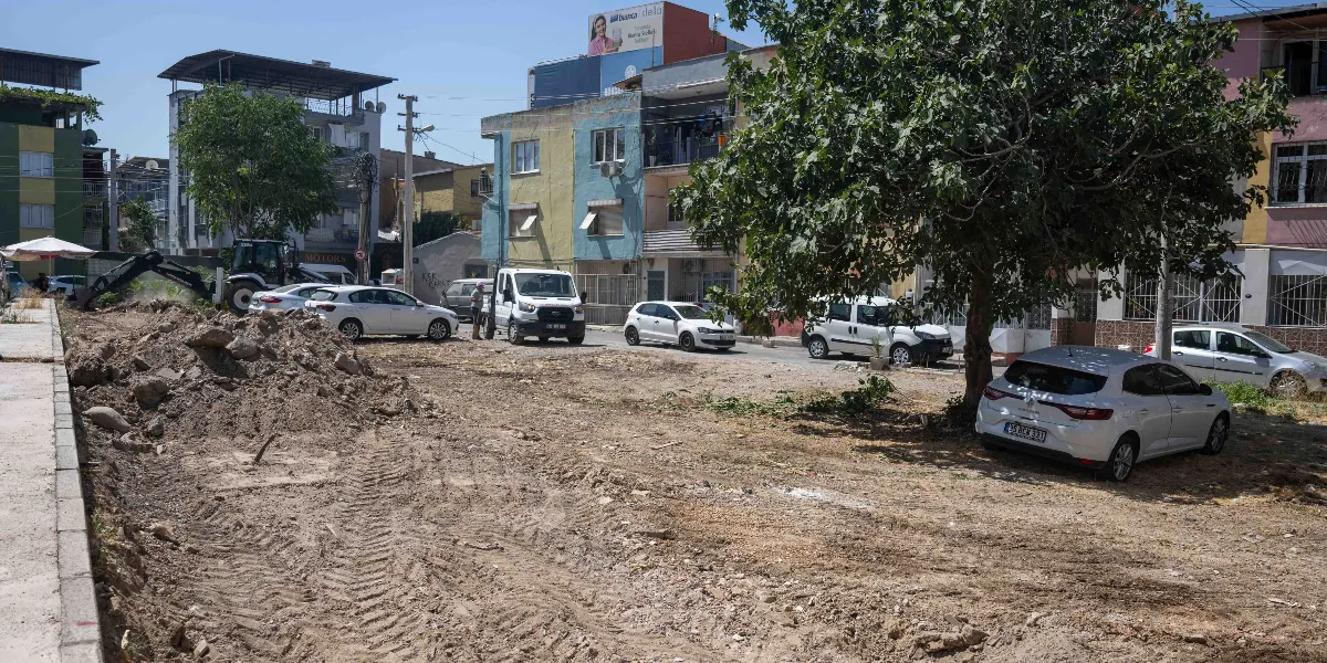 Bornova'da toprak zemin üzerinde devam eden çevre düzenleme çalışmaları görülüyor. Sol tarafta bir iş makinesi kazı yaparken, sağ tarafta büyük bir ağacın gölgesinde park etmiş beyaz bir otomobil bulunuyor. Arka planda ise çok katlı apartmanlar, park halindeki diğer araçlar ve açık bir gökyüzü yer alıyor.