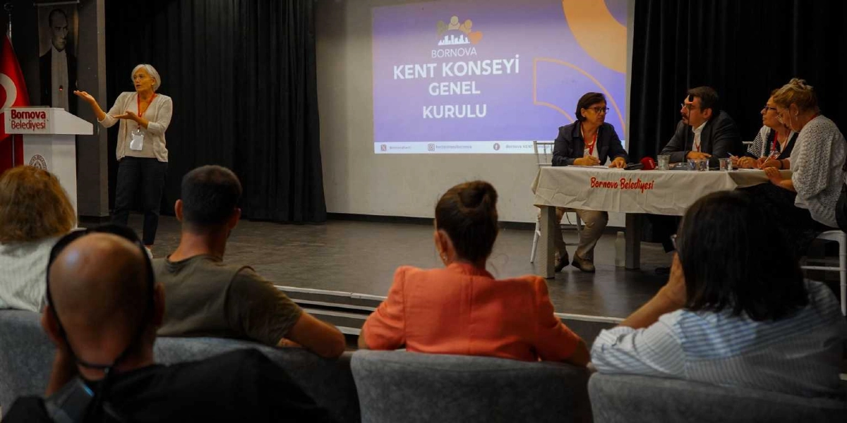 Bornova Kent Konseyi Genel Kurulu toplantısında, kürsüde konuşma yapan bir kadın ve masada oturan dört kişilik bir kurul heyeti izleyicilere hitap ediyor. Salonun ön tarafındaki dev ekranda Bornova Kent Konseyi logosu ve Genel Kurulu yazısı yer alırken, izleyiciler koltuklarından toplantıyı takip ediyor. Kürsünün üzerinde Bornova Belediyesi logosu bulunuyor.