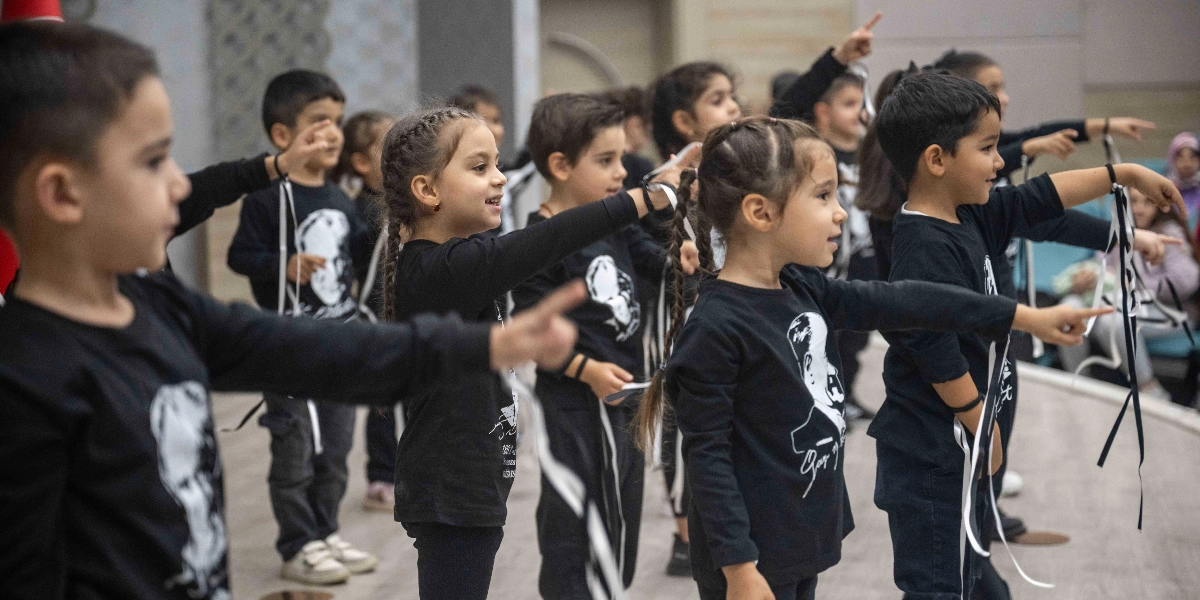 Üzerinde Atatürk silüeti ve imzası bulunan siyah tişörtler giymiş bir grup anaokulu öğrencisi, bir gösteri veya anma etkinliği kapsamında aynı yöne doğru ellerini uzatarak duruyor. Çocukların bileklerine siyah kurdeleler bağlanmış durumda ve hepsi ciddiyetle ileriye odaklanmış görünüyor.