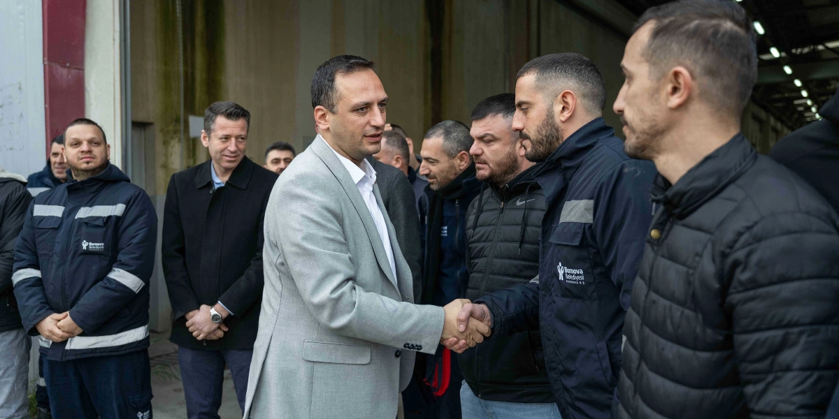 Açık gri ceketli ve beyaz gömlekli Başkan Ömer Eşki, üzerinde Bornova Belediyesi Personel A.Ş. logolu mont bulunan bir çalışanla el sıkışıyor. Çevredeki diğer çalışanlar ve yetkililer, kapalı bir depo veya atölye ortamında onları izliyor.