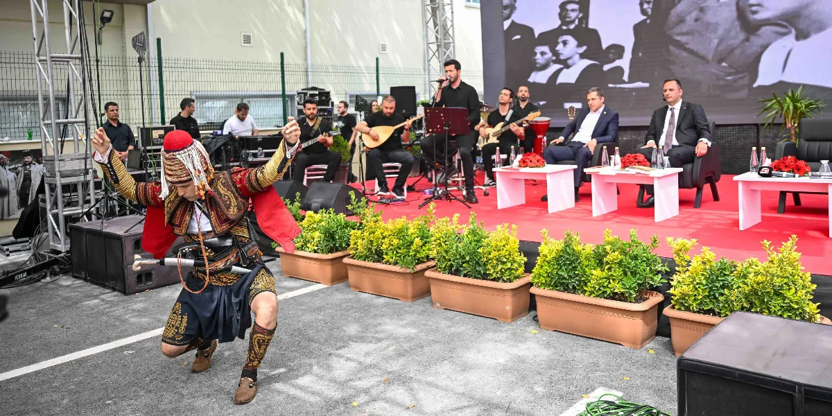 Açık havada düzenlenen bir etkinlikte, geleneksel zeybek kıyafetleri giymiş bir dansçı diz çökmüş figür sergiliyor. Arka planda kırmızı halı serili sahnede müzisyenler enstrümanlarıyla performans sergiliyor, yanlarında ise takım elbiseli iki kişi masada oturuyor. Sahnenin arkasındaki dev ekranda siyah beyaz tarihi fotoğraflar yer alıyor.