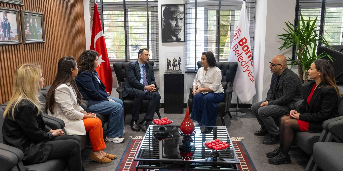 Bornova Belediye Başkanı ve beraberindeki heyet, bir ofis ortamında karşılıklı oturarak toplantı yapıyor. Odanın bir köşesinde Türk bayrağı ve Bornova Belediyesi bayrağı yer alırken, duvarda Mustafa Kemal Atatürk'ün portresi asılı. Ortadaki cam sehpanın üzerinde kırmızı dekoratif objeler ve ikramlar bulunuyor. Katılımcılar resmi ve yarı resmi kıyafetlerle bir araya gelerek görüşme gerçekleştiriyor.