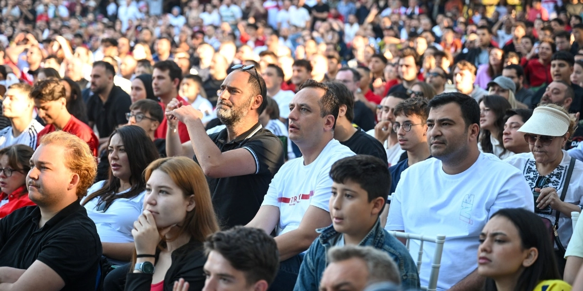 Açık havada düzenlenen bir etkinlikte, sahneye veya ekrana odaklanmış, farklı yaş gruplarından oluşan kalabalık bir izleyici grubu. Ön planda, beyaz tişörtlü ve dikkatli bir şekilde ileriye bakan orta yaşlı bir erkek ile yanında oturan kot ceketli bir çocuk dikkat çekiyor. İzleyicilerin çoğu gündelik kıyafetler içinde, bazıları güneş gözlüğü takıyor ve genel olarak ciddi veya meraklı bir ifadeyle etkinliği takip ediyorlar.