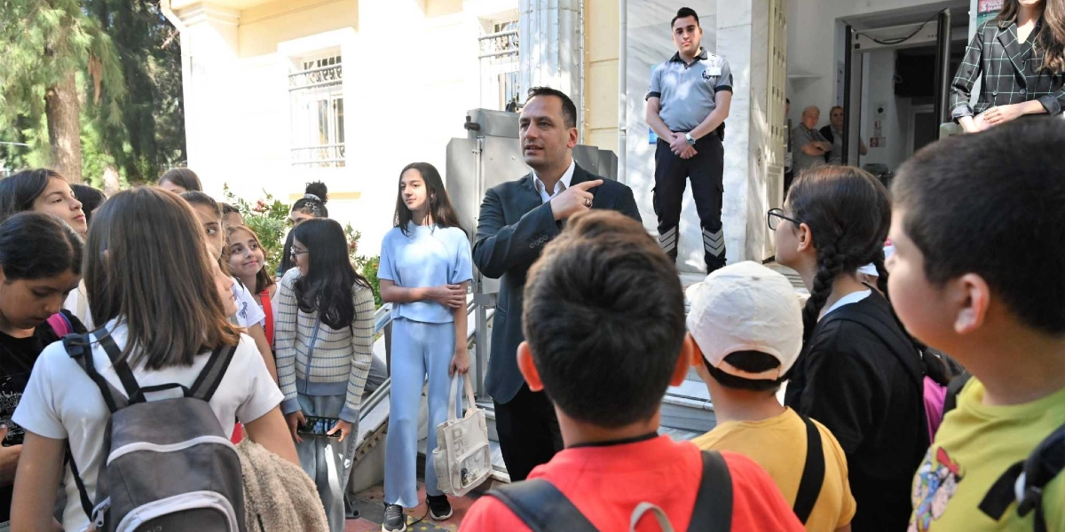 Bornova Belediyesi binasının önünde, takım elbiseli bir yetkili, etrafını saran ilkokul ve ortaokul çağındaki bir grup öğrenciye bir şeyler anlatarak binayı işaret ediyor. Öğrenciler dikkatle dinlerken, arkada üniformalı bir güvenlik görevlisi bekliyor.