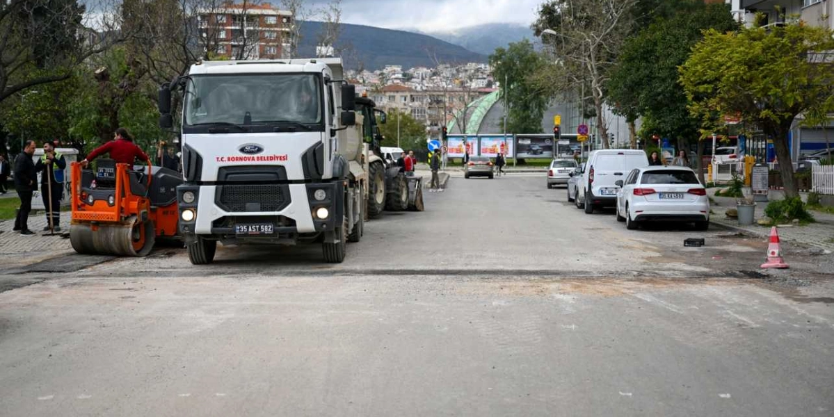 Bornova Belediyesi'ne ait beyaz bir damperli kamyon ve turuncu bir silindir, şehir merkezindeki bir caddede yol yenileme çalışması yapıyor. Çalışma alanının yanında işçiler bulunurken, caddenin sağ tarafında park halindeki araçlar ve arka planda ağaçlıklı bir yerleşim bölgesi görülüyor.