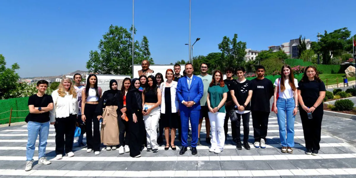 Açık havada, güneşli bir günde, taş döşeli bir meydanda yan yana duran kalabalık bir öğrenci grubu ve ortalarında mavi takım elbiseli bir erkek ile beyaz önlüklü bir kadının yer aldığı toplu fotoğraf. Arka planda ağaçlar, binalar ve bir büst bulunmaktadır.