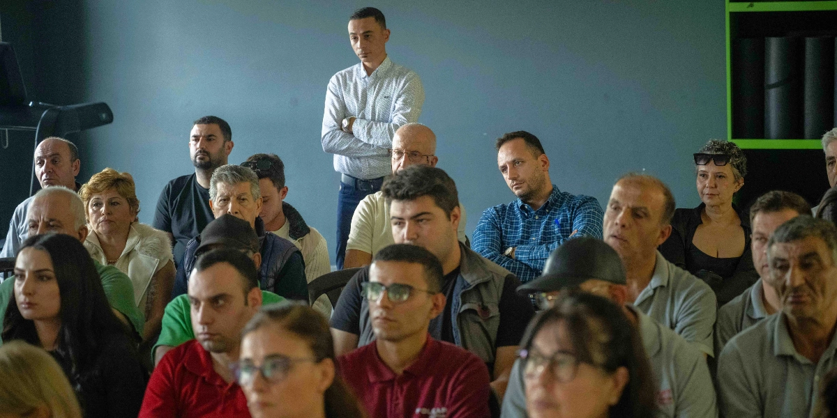 Kapalı bir alanda, bir sunumu veya konuşmayı dikkatle dinleyen, farklı yaş gruplarından oluşan kalabalık bir katılımcı grubu. Arka planda ayakta duran bir kişi ve ön sıralarda oturan, çeşitli renklerde kıyafetler giymiş, gözlüklü ve gözlüksüz kadın ve erkeklerden oluşan izleyiciler.
