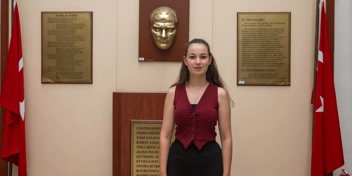 Koyu kırmızı yelek ve siyah pantolon giymiş genç bir kadın, arka planda Mustafa Kemal Atatürk'ün kabartma büstü, iki yanında Türk bayrakları ve duvarda İstiklal Marşı ile Gençliğe Hitabe metinlerinin yer aldığı resmi bir ortamda gülümseyerek duruyor.