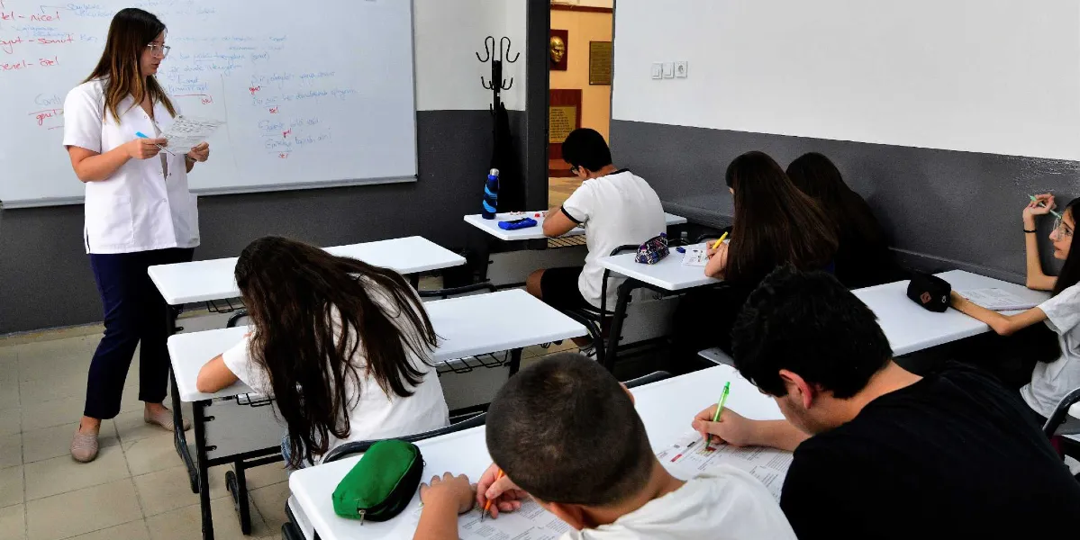 Sınıf ortamında beyaz önlüklü bir öğretmen, elindeki kağıdı inceleyerek tahtanın önünde duruyor. Öğrenciler sıralarında oturmuş, önlerindeki test kağıtlarını dikkatle çözüyor. Tahtada dil bilgisi konularıyla ilgili el yazısı notlar yer alıyor.