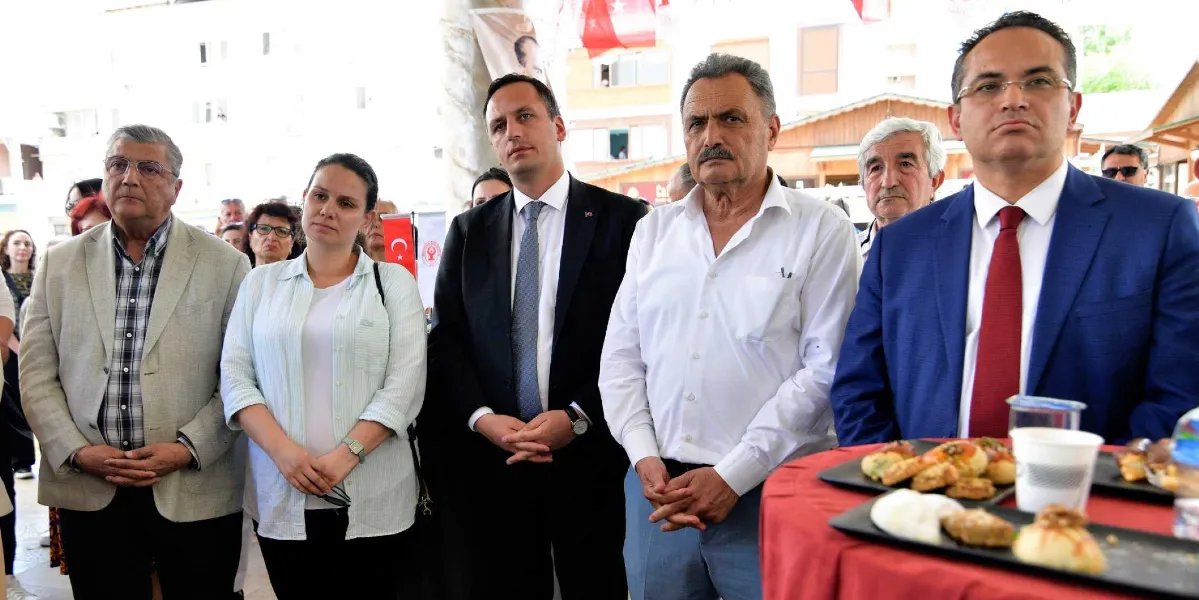 Açık havada düzenlenen bir etkinlikte yan yana duran, aralarında Bornova Belediye Başkanı Mustafa İduğ’un da bulunduğu bir grup bürokrat ve yetkili. Ön planda, üzerinde çeşitli atıştırmalıkların bulunduğu kırmızı örtülü bir masa yer alıyor. Katılımcılar ciddi bir ifadeyle karşıya bakarken, arka planda Türk bayrakları ve belediyeye ait görseller seçiliyor.